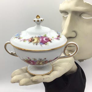 May include: A white porcelain lidded serving bowl with floral designs in pink, purple, yellow, and blue. The bowl has gold trim and handles. The lid is topped with a gold finial. The bowl is held by a statue of a hand.