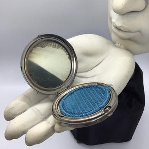 May include: An open, antique silver-toned compact mirror with a blue powder puff. The mirror is round and the compact is oval. The compact is held by a white hand sculpture against a black background.