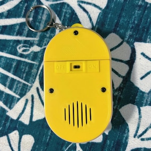 May include: A yellow, oval-shaped keychain accessory with a silver key ring. The accessory has an on/off switch and a speaker-like design with vertical lines. The word "OFF" and "ON" are printed on the accessory.
