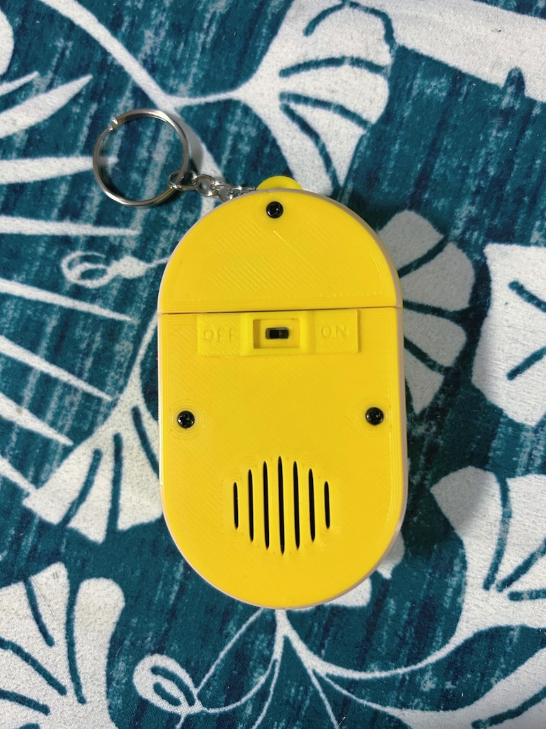 May include: A yellow, oval-shaped keychain accessory with a silver key ring. The accessory has an on/off switch and a speaker-like design with vertical lines. The word "OFF" and "ON" are printed on the accessory.
