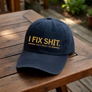 May include: Navy blue baseball cap with the embroidered text "I FIX SHIT. THAT'S WHAT I DO" in gold thread. The cap is sitting on a wooden surface.
