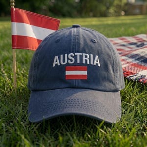May include: A navy blue baseball cap featuring the word AUSTRIA embroidered in white capital letters above a small, rectangular red and white Austrian flag emblem centered on the front panel of the hat, which has a curved brim and stitched eyelets.