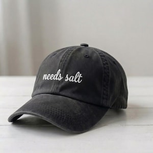 May include: A charcoal grey baseball cap with a curved brim and a button on top. The front of the cap features the text "needs salt" embroidered in white cursive script. The fabric has a slightly faded, washed-out appearance throughout the design.