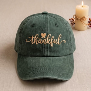 May include: A dark green baseball cap with a distressed fabric finish. The front features the word thankful embroidered in a light tan cursive script, with a small heart icon positioned directly above the letter k.
