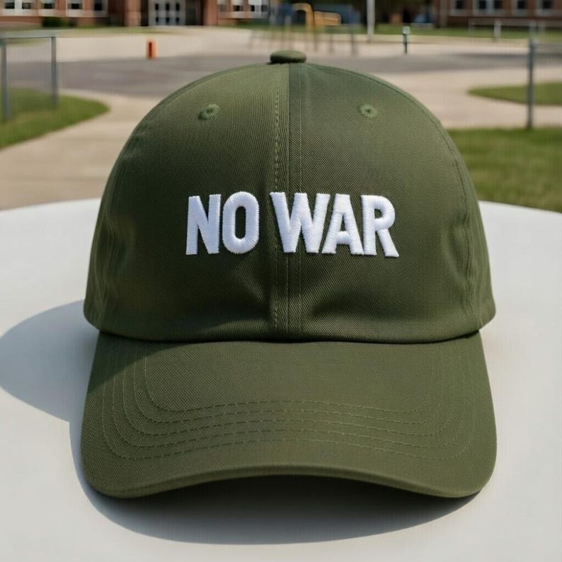 May include: Olive green baseball cap with the white embroidered text "NO WAR" across the front. The cap has a curved brim and a button on top. The background shows a schoolyard.