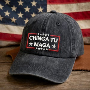 May include: A dark gray baseball cap with the embroidered text "CHINGA TU MAGA" in white letters inside a red rectangle, with white stars above and below. The background includes an American flag.