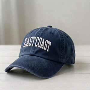 Puede incluir: Una gorra de b&eacute;isbol azul marino con un acabado desgastado de estilo vintage. La parte frontal de la gorra presenta el texto EAST COAST bordado en letras may&uacute;sculas blancas con una fuente arqueada. La gorra tiene una corona estructurada.