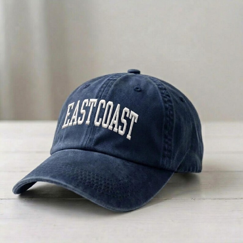 Puede incluir: Una gorra de b&eacute;isbol azul marino con un acabado desgastado de estilo vintage. La parte frontal de la gorra presenta el texto EAST COAST bordado en letras may&uacute;sculas blancas con una fuente arqueada. La gorra tiene una corona estructurada.