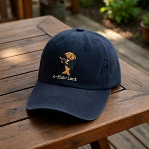 May include: Navy blue baseball cap with an embroidered design of a fish wearing a tuxedo and holding a book. The text "o-fish-iant" is embroidered below the fish. The cap is sitting on a wooden table.