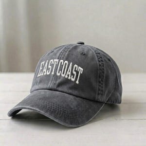 Puede incluir: Una gorra de b&eacute;isbol gris carb&oacute;n con un acabado desgastado y lavado. La parte frontal presenta el texto EAST COAST bordado en letras may&uacute;sculas blancas. La gorra tiene una visera curva, una corona estructurada con ojales y un bot&oacute;n superior.