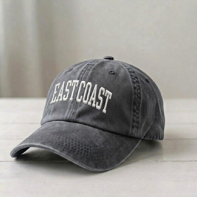 Puede incluir: Una gorra de b&eacute;isbol gris carb&oacute;n con un acabado desgastado y lavado. La parte frontal presenta el texto EAST COAST bordado en letras may&uacute;sculas blancas. La gorra tiene una visera curva, una corona estructurada con ojales y un bot&oacute;n superior.