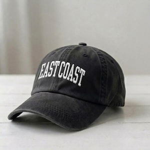 Puede incluir: Una gorra de b&eacute;isbol gris carb&oacute;n con el texto EAST COAST bordado en letras may&uacute;sculas blancas en el panel frontal. La gorra tiene una visera curva, un bot&oacute;n en la parte superior de la corona y un dise&ntilde;o estructurado con costuras visibles.
