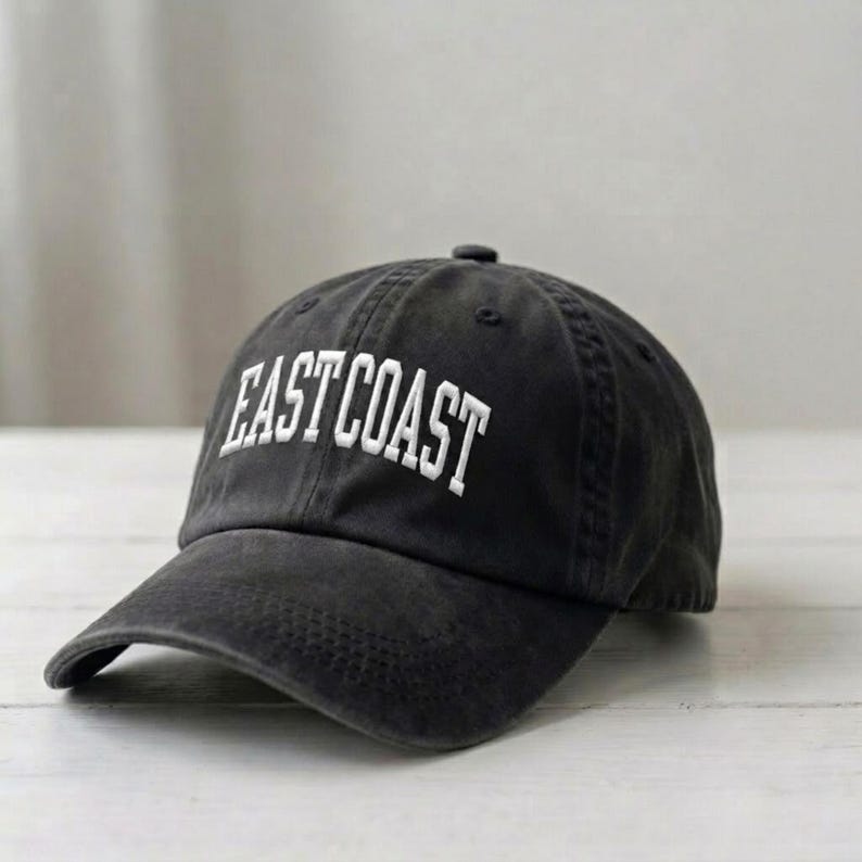 Puede incluir: Una gorra de b&eacute;isbol gris carb&oacute;n con el texto EAST COAST bordado en letras may&uacute;sculas blancas en el panel frontal. La gorra tiene una visera curva, un bot&oacute;n en la parte superior de la corona y un dise&ntilde;o estructurado con costuras visibles.