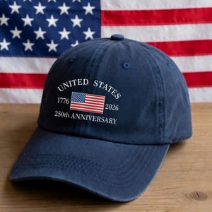 May include: A navy blue baseball cap featuring white embroidered text that reads UNITED STATES, 1776, 2026, and 250th ANNIVERSARY, centered around a small, rectangular, embroidered American flag graphic on the front panel of the cap.