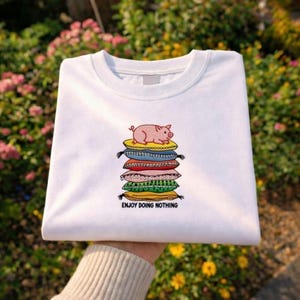 May include: White crew neck sweatshirt with an embroidered design of a pink pig on a stack of colorful pillows. The text "ENJOY DOING NOTHING" is below the design.