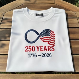 May include: White t-shirt featuring a navy blue infinity symbol intertwined with a waving American flag. The text "250 YEARS 1776-2026" is embroidered in red and navy blue. The shirt is folded neatly.