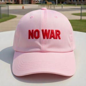 May include: A light pink baseball cap with the embroidered text "NO WAR" in red. The cap has a curved brim and a rounded top with a small button. The background shows a blurred outdoor scene.