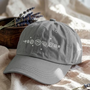 May include: A gray baseball cap featuring a white embroidered design of the planets in our solar system. The cap has a curved brim and a button on top. The design includes the sun, Mercury, Venus, Earth, Mars, Jupiter, Saturn, Uranus, and Neptune.
