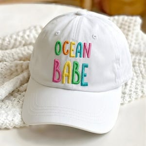 May include: White baseball cap with the embroidered text "OCEAN BABE" in rainbow colors. The hat has a curved brim and a button on top. The hat is made of a soft material.