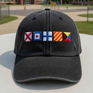 May include: A dark gray baseball cap with embroidered nautical signal flags in red, white, blue, and yellow. The cap has a curved brim and a fabric strap closure. The hat is made of a soft, worn material.