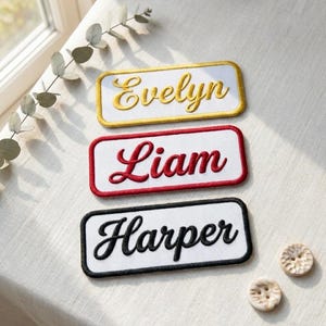 Puede incluir: Tres parches bordados con nombres: "Evelyn" en dorado sobre un borde amarillo, "Liam" en rojo sobre un borde rojo y "Harper" en negro sobre un borde negro. Los parches son rectangulares con esquinas redondeadas, sobre una superficie clara.