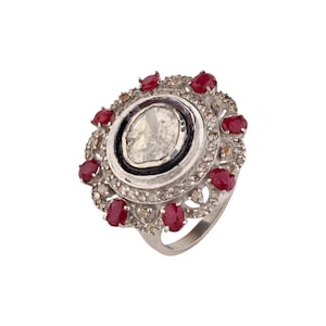 以下が含まれることがあります： 中央に不規則な形の透明な石を配し、黒い縁で囲まれたシルバーリング。リングは、小さな丸い赤い宝石と透明な石で装飾され、花のようなデザインを作り出しています。豪華なジュエリーです。