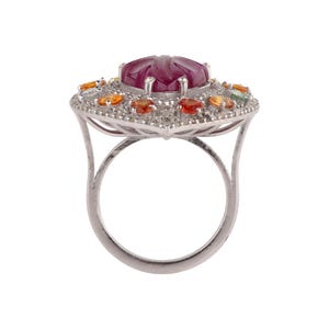 以下が含まれることがあります： 中央に大きく彫刻された楕円形の濃い赤色の宝石をあしらったシルバーリング。周囲には小さくカラフルな宝石が散りばめられています。リングは、詳細な透かし彫りのデザインと研磨仕上げが施されています。