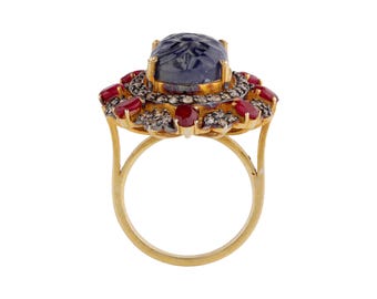 May include: A gold ring with a large, oval, blue gemstone with an engraved design. The gemstone is encircled by small red gemstones and clear stones. The ring has a delicate, openwork design, typical of fine jewellery.