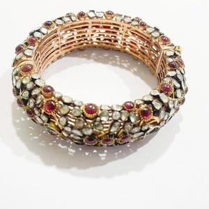 May include: A gold-toned bangle bracelet with a detailed design. The bracelet features numerous small, clear stones and vibrant red gemstones. The inner structure of the bracelet is visible, showcasing a layered, openwork design. The bracelet is set against a white background.