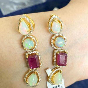 Può includere: Un paio di orecchini d'oro con opali a goccia, opali rotondi e rubini rettangolari. Le pietre preziose sono incastonate con piccoli diamanti. Un accessorio di lusso.