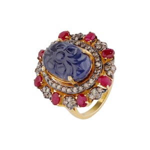 May include: A gold ring with a central, carved blue gemstone, encircled by red gemstones and a halo of smaller stones. The ring showcases an intricate, ornate design.