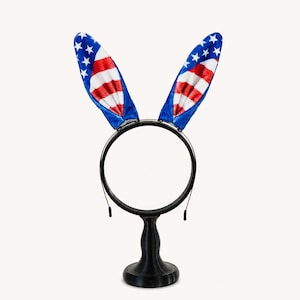 Peut inclure: Un serre-tête noir doté de deux oreilles de lapin dressées, décorées du motif du drapeau américain avec des étoiles blanches sur fond bleu et des rayures horizontales rouges et blanches. Le serre-tête est posé sur un support noir.