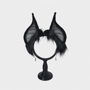 May include: A black headband featuring two tall, pointed bat-like ears. The ears are constructed from a thin, semi-translucent fabric with visible structural lines, surrounded by long, shaggy black faux fur at the base of each ear.
