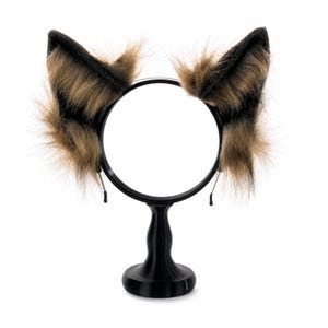 May include: A pair of faux fur animal ears attached to a black circular headband. The ears feature a mix of dark brown and tan long-pile synthetic fur, with a pointed shape and a textured appearance, mounted on a black pedestal display stand.