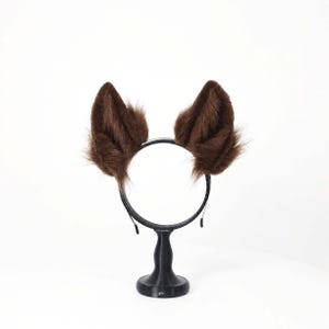 May include: A headband featuring two upright, pointed animal ears covered in long, dark brown faux fur. The ears are attached to a thin, black circular band, creating a structured accessory that mimics the appearance of a fox or wolf.