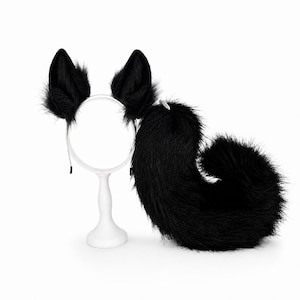 May include: A set consisting of a black faux fur headband with two pointed animal-like ears and a matching long, thick, black faux fur tail. The headband is positioned on a white stand, while the curved tail rests beside it on a white surface.