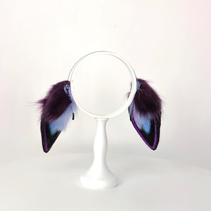 May include: A pair of faux fur cosplay ears in shades of purple, black, and blue, attached to a white headband. The ears are displayed on a white stand against a white background. The ears have a fluffy texture.