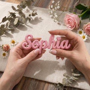 May include: A pink embroidered patch spelling "Sophia" in a cursive font, held by hands. The patch features a heart detail above the letter "a". The background includes flowers and foliage.