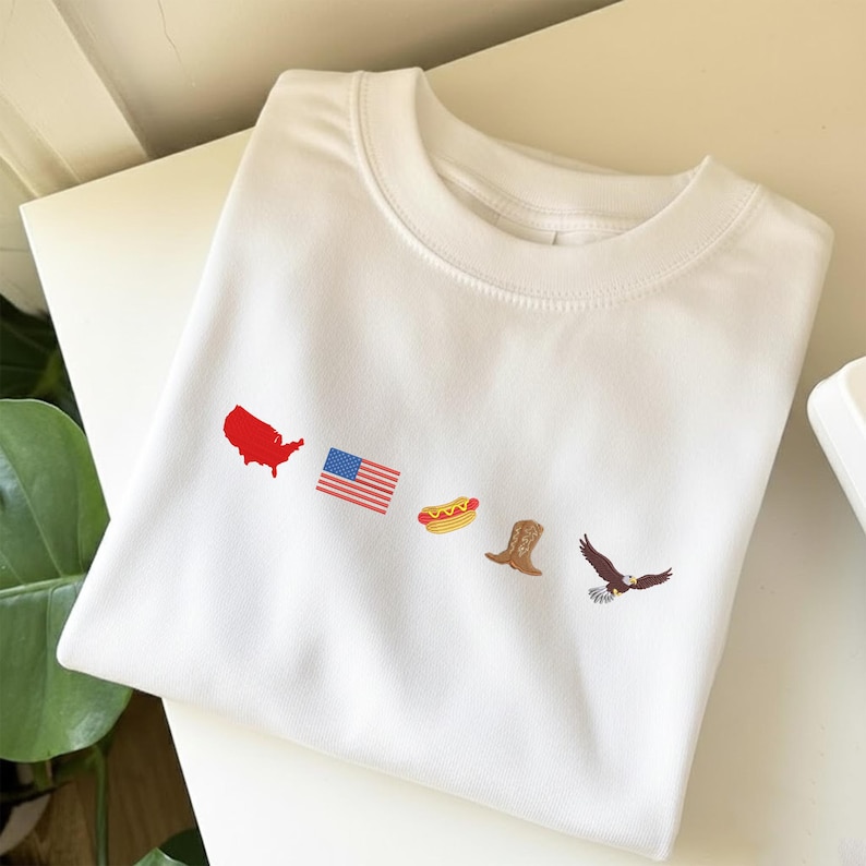 K&ouml;nnte beinhalten: Wei&szlig;es Sweatshirt mit Rundhalsausschnitt und gestickten patriotischen Symbolen. Das Design umfasst eine rote Umrisskarte der Vereinigten Staaten, eine US-Flagge, einen Hotdog, einen Cowboy-Stiefel und einen braunen Adler mit ausgebreiteten Fl&uuml;geln.