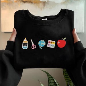 May include: Black sweatshirt with school-themed patches. Patches include a glue bottle, scissors, a globe, a palette of colorful faces, a hashtag, and a red apple. The sweatshirt has a crew neck and a distressed design.