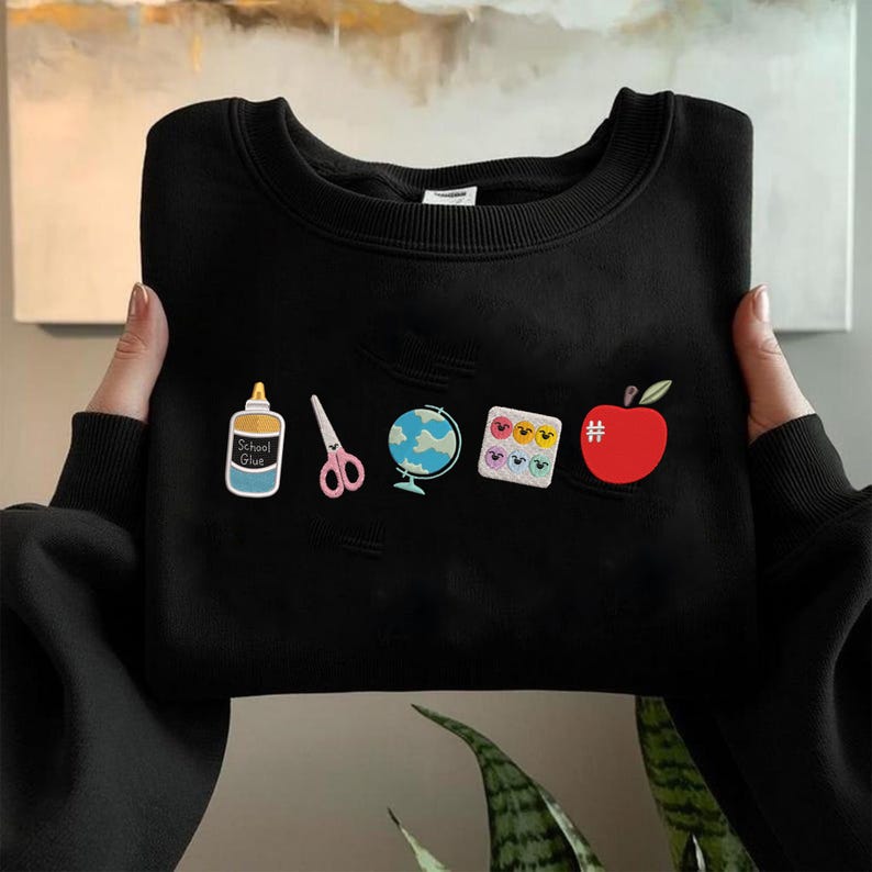 May include: Black sweatshirt with school-themed patches. Patches include a glue bottle, scissors, a globe, a palette of colorful faces, a hashtag, and a red apple. The sweatshirt has a crew neck and a distressed design.