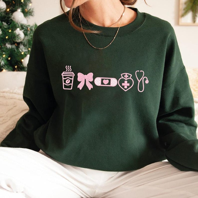 May include: Dark green sweatshirt with pink graphic icons including a coffee cup, bow, bandage, heart with a cross, and a stethoscope. The sweatshirt is a crew neck style and appears to be made of a soft material.