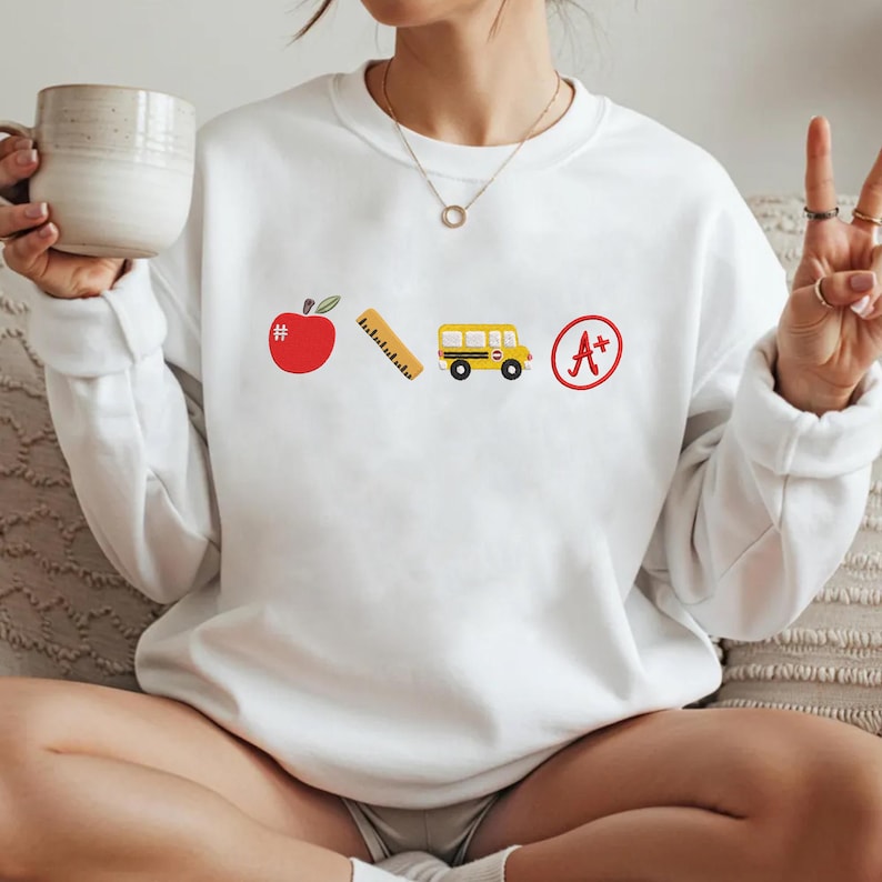 May include: White sweatshirt with colorful school-themed graphics. The design includes a red apple with a hashtag, a ruler, a yellow school bus, and a red A+ grade symbol. The sweatshirt is a casual, comfortable style.