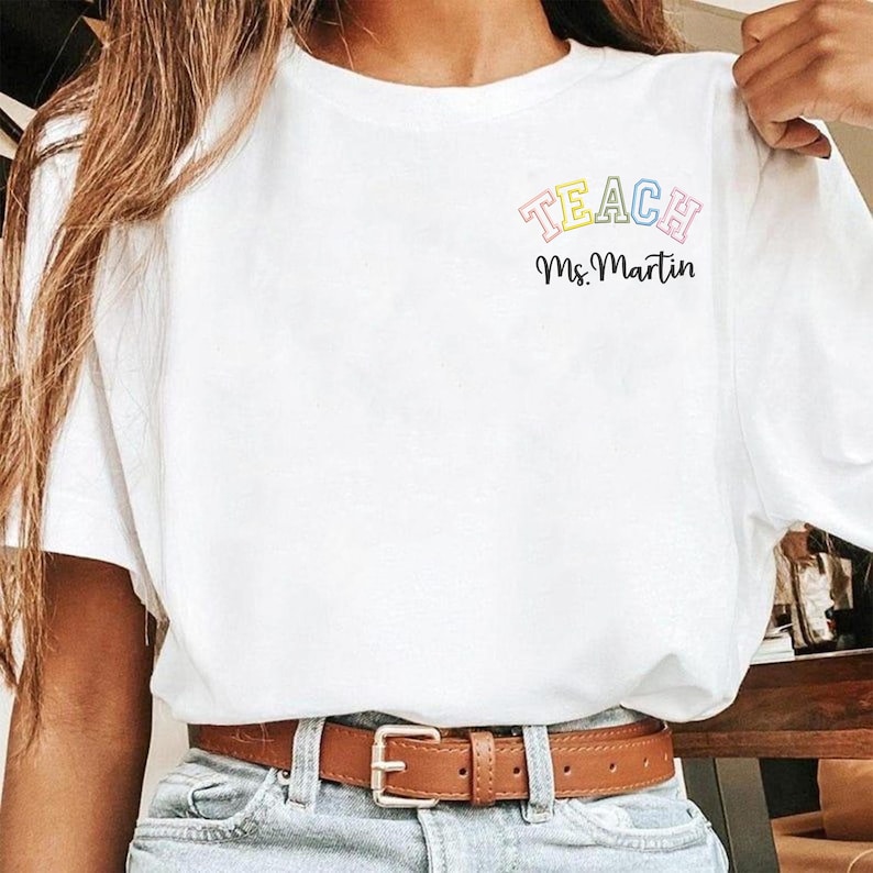 May include: A white t-shirt with the word "TEACH" in pastel colors and the name "Ms. Martin" in black script. The shirt is paired with blue jeans and a brown belt.