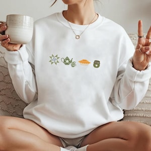 K&ouml;nnte beinhalten: Wei&szlig;es Sweatshirt mit gestickten Abbildungen eines Quiltblocks, einer Teekanne, einer Teetasse, einem Kuchen und einem Glas. Das Design ist in Gr&uuml;n-, Gelb- und Braunt&ouml;nen gehalten. Das Sweatshirt ist ein l&auml;ssiges, bequemes Kleidungsst&uuml;ck.