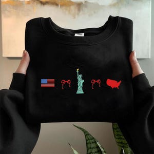 K&ouml;nnte beinhalten: Schwarzes Sweatshirt mit einem Used-Look-Design mit patriotischen Grafiken. Das Design umfasst die amerikanische Flagge, eine Schleife, die Freiheitsstatue, eine weitere Schleife und eine rote Umrisskarte der Vereinigten Staaten.