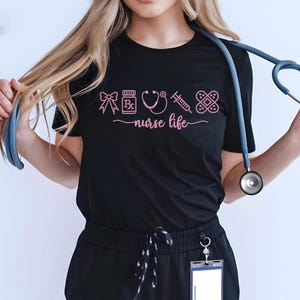 May include: Black t-shirt with pink graphic icons of medical supplies and the words "nurse life". A stethoscope hangs around the neck. The shirt is made of a soft material.