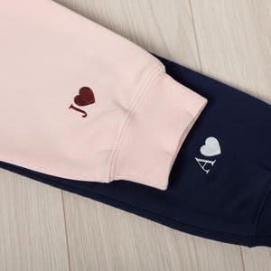 Op de afbeelding: Close-up van een roze sweatshirtmouw met een bordeauxrood hart en de letter "J" geborduurd. Daaronder een marineblauwe joggingbroekmouw met een wit hart en de letter "A". De kleding is zacht en casual.