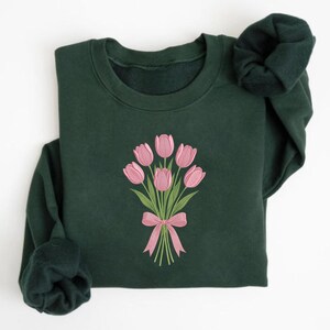 K&ouml;nnte beinhalten: Ein dunkelgr&uuml;nes Sweatshirt mit einem gestickten Bouquet aus rosa Tulpen, das mit einer rosa Schleife gebunden ist. Die langen &Auml;rmel sind gefaltet und zeigen den weichen Stoff und das Design des Sweatshirts.