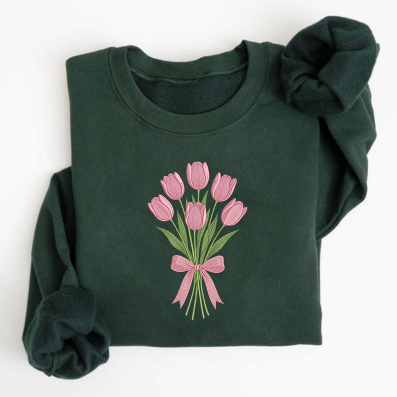 K&ouml;nnte beinhalten: Ein dunkelgr&uuml;nes Sweatshirt mit einem gestickten Bouquet aus rosa Tulpen, das mit einer rosa Schleife gebunden ist. Die langen &Auml;rmel sind gefaltet und zeigen den weichen Stoff und das Design des Sweatshirts.